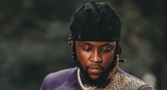 DJ ShabZi Madallion, with a beard and braided hair, wearing a black cap, purple jacket, and a chain necklace, looks down with a neutral expression against a blurred background—ready for epic hip hop nights and freestyles.