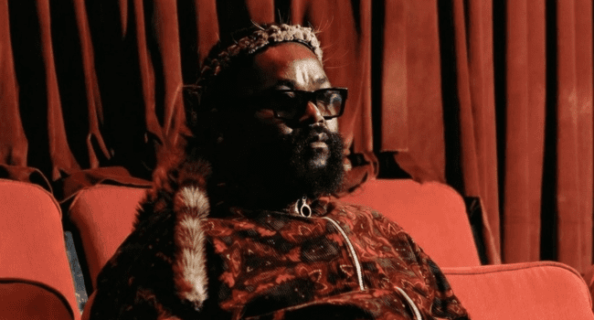 A person with a beard and glasses sits on a red sofa, wearing a patterned garment and headpiece, with red curtains in the background—a scene reminiscent of Sjava’s iNkanyezi NeZinkanyezi album cover.