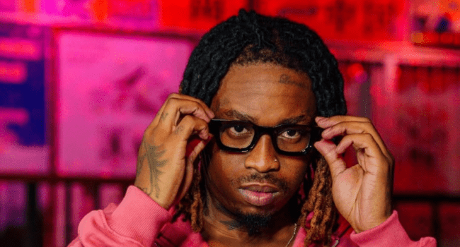 A person with dreadlocks and tattoos adjusts black glasses while looking forward in a pink top, channeling Valentine’s vibes against a colorful, blurred background.