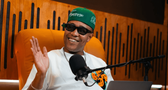 A person wearing sunglasses and a green cap sits in front of a microphone, smiling and gesturing with one hand in a studio setting, embodying DJ Speedsta's intentional approach during the Up To Speed podcast.