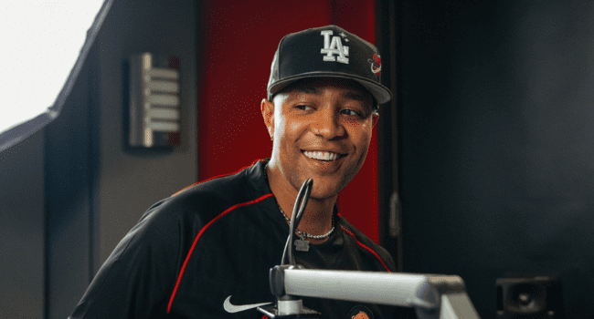 A man wearing a black LA baseball cap and a black sports shirt smiles while standing in front of a microphone in a recording studio, channeling DJ Speedsta vibes as he works on tracks like "Special Somebody.