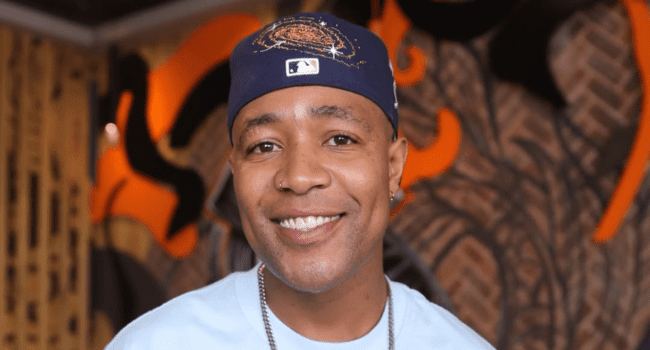 A person wearing a blue baseball cap and light blue shirt smiles at the camera, with abstract art on the wall in the background, capturing the upbeat vibe of SA Hip-Hop.