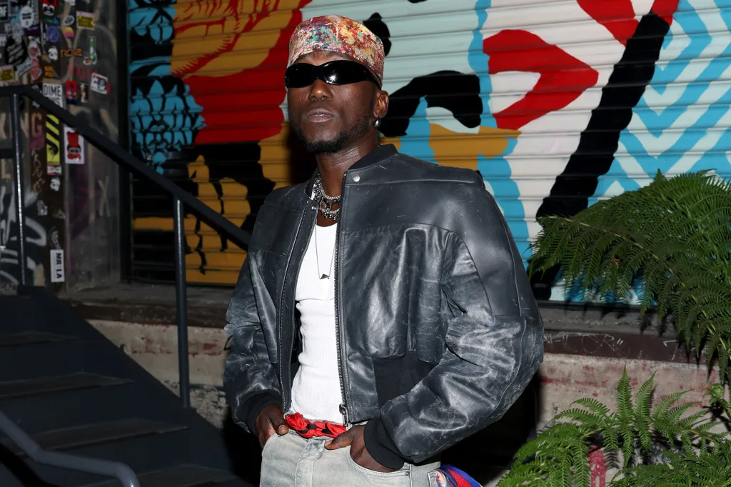 A person wearing a patterned headscarf, dark sunglasses, a black leather jacket, and white shirt stands in front of a colorful mural, channeling the bold style seen on African Fresh Picks from billboard.com.