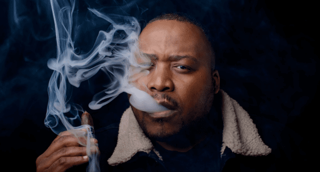 A man in a dark jacket holds a cigar and exhales smoke, which swirls in front of his face against a dark background, evoking the moody vibe of a Stogie T music video.
