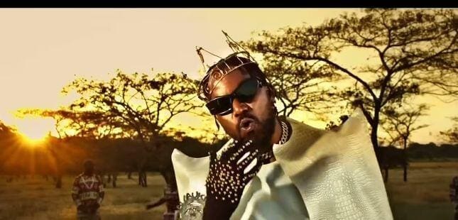 A man wearing a gold crown, sunglasses, and a dramatic white and gold outfit stands outdoors at sunset—channeling Tay Grin’s iconic comeback style—with trees and other people visible in the background.