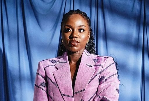 A woman wearing a pink blazer stands in front of a blue curtain, looking slightly to the side with a neutral expression—an image capturing the confident poise of Billboard 2025’s Global Power Players in African music.