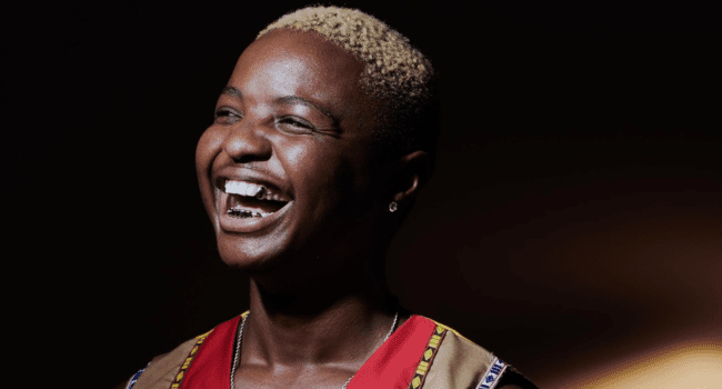 A person with short blond hair and a patterned vest smiles broadly against a dark background, capturing the spirit of Tony Dayimane and celebrating musical growth.
