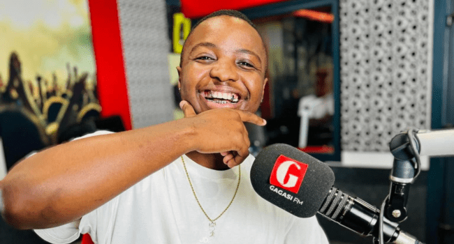 A man smiling and gesturing with his hand while seated at a radio studio microphone labeled "Gagasi FM," chats about Tony Dayimane's latest EP, "Girls & MP3’s.