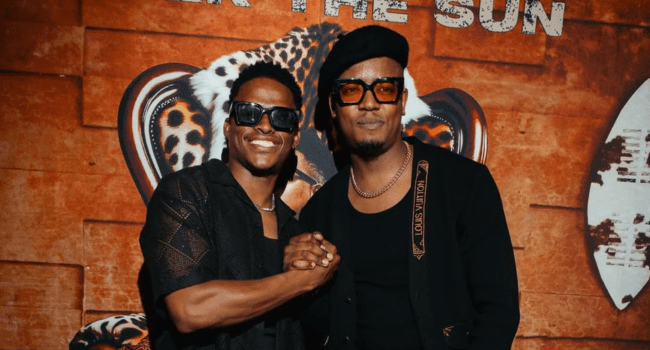 Two men wearing black outfits and sunglasses shake hands and smile in front of a leopard graphic backdrop, capturing the vibrant energy of Sun-El Musician’s "Under The Sun.