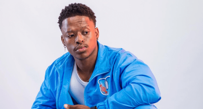 A man wearing a blue jacket over a white shirt sits against a plain light background, looking slightly to his left with a neutral expression, reminiscent of Touchline on the cover of his acclaimed album S.O.O.N 2.