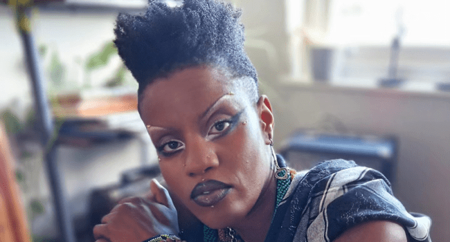 Person with bold makeup and short, styled hair sits indoors, looking directly at the camera. Shelves and a window with light are visible in the blurred background, evoking an edgy vibe inspired by Toya Delazy’s journey in the SA music industry.