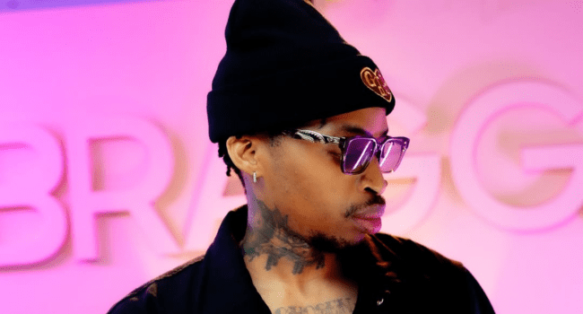A person wearing a black beanie, sunglasses, and a black jacket stands in front of a pink and white background with large, blurred letters—perfect for a Tshego or Podcast and Chill vibe.