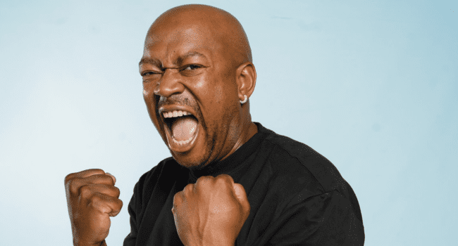 Bald man in a black shirt with clenched fists, mouth wide open and an intense expression, standing against a light blue background—capturing the energy of the Tswyza Show’s favourite episodes.