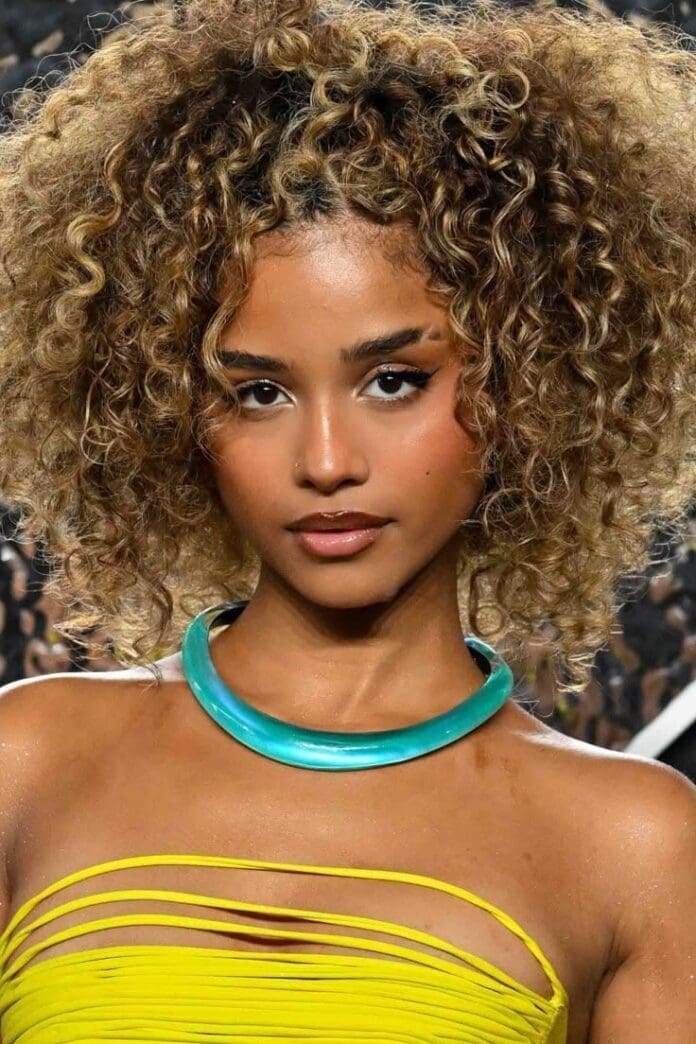 A woman with curly blonde hair wears a yellow top with cutouts and a chunky turquoise necklace, channeling Tyla’s Grammy win style as she poses against a blurred background.
