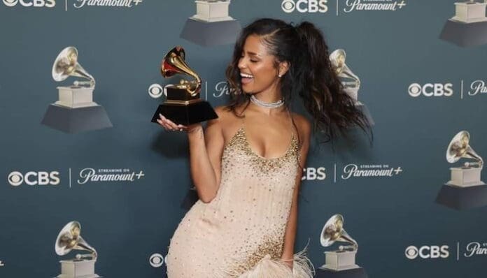 Tyla, in a sparkly dress, smiles and holds a Grammy award for Best African Music Performance in front of a backdrop with Grammy, CBS, and Paramount+ logos at the Grammys 2026.