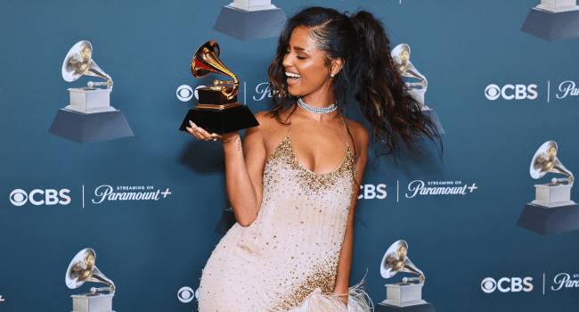 Tyla, in a sparkly dress, holds a Grammy award and smiles on a blue step-and-repeat backdrop with CBS and Paramount+ logos, celebrating as one of the Africans to win Grammys.