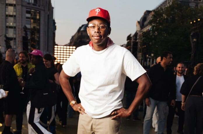 A man in a white T-shirt, red cap, and glasses stands outdoors with hands on hips, surrounded by people at an event featuring lively rap songs and African music samples.