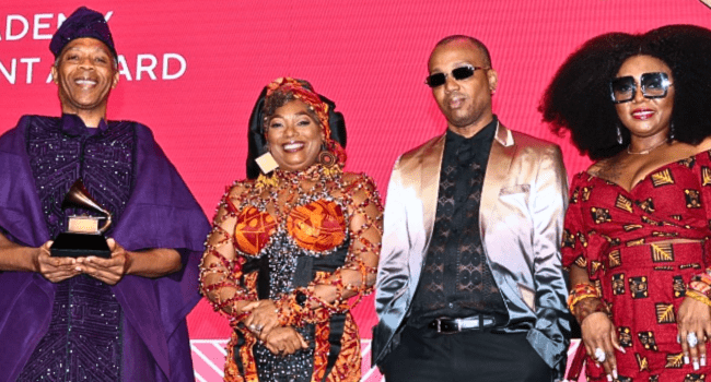 Untitled-design-2.png Four people in colorful formal attire pose together at an awards event; one holds a Grammy trophy, celebrating Fela Kuti's legacy, in front of a red background with partial event signage.