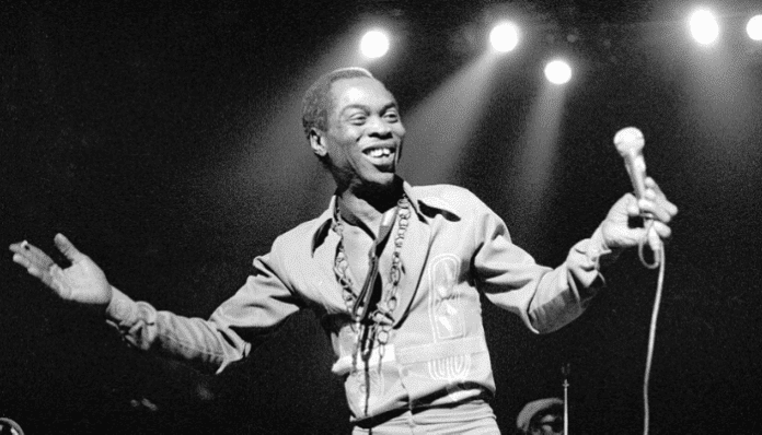 A man in a patterned shirt smiles on stage while holding a microphone, with stage lights shining in the background, echoing the vibrant spirit of Africa and icons like Fela Kuti.