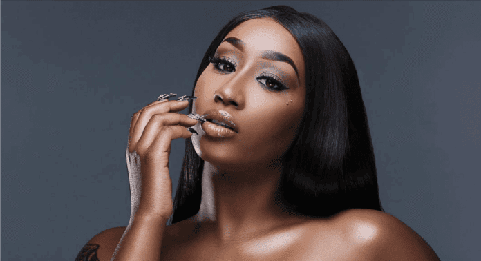 A woman with long straight black hair and glossy lips poses against a plain dark background, touching her face with manicured nails—a look reminiscent of Victoria Kimani’s signature style.