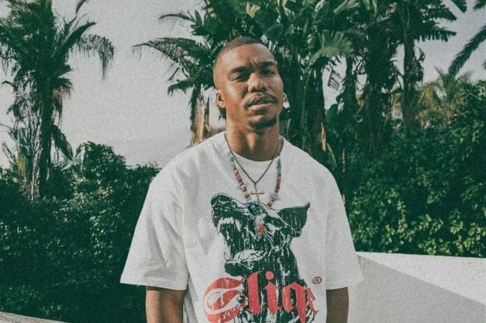 A man stands outdoors in front of palm trees, wearing a white graphic t-shirt featuring a dog's face, layered necklaces, and looking toward the camera—channeling DJ Sliqe’s signature Hip Hop style.