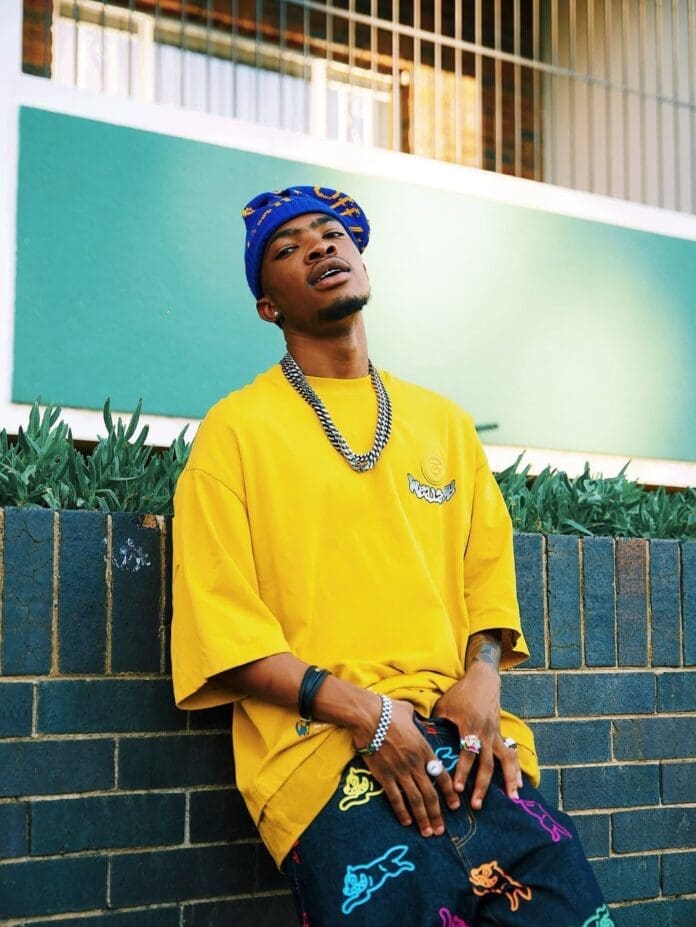 A person wearing a yellow shirt, blue beanie, and colorful pants sits against a brick wall, exuding cool Royal MusiQ vibes, with greenery and a building in the background.