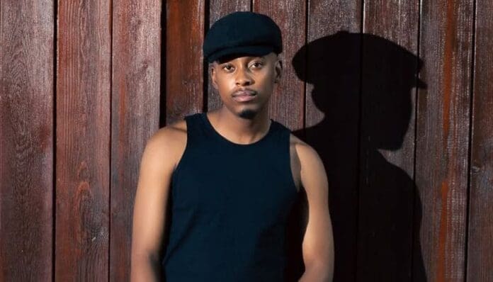 A man wearing a black cap and black sleeveless shirt stands against a wooden wall with his shadow cast beside him, channeling the vibe of an album release featured on SlikourOnLife.