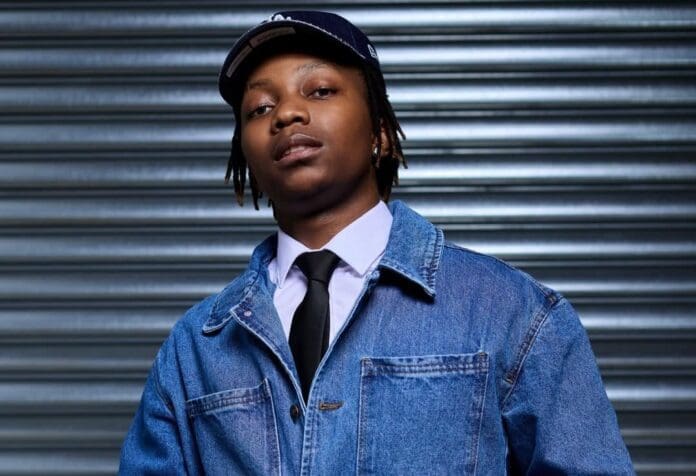 A person wearing a black cap, denim jacket, white shirt, and black tie stands in front of a metallic roll-up door, channeling DJ Maphorisa’s cool and confident vibe.