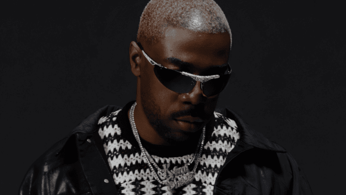 A man with short, bleached hair wears dark sunglasses, a patterned sweater, layered silver chains, and a black jacket against a dark background, channeling the striking visuals often seen in James BKS or Milli Vanity’s signature styles.