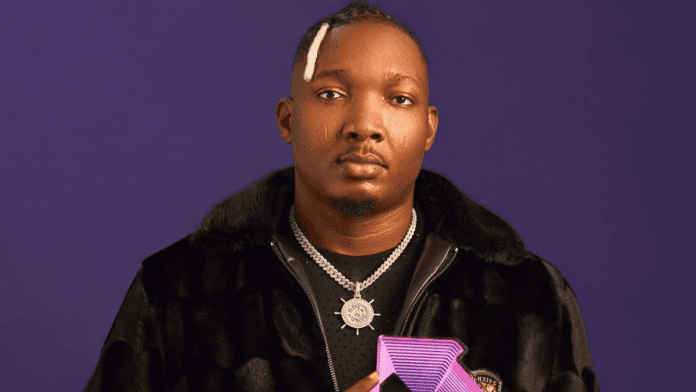 A man in a black fur-collared jacket and chain necklace stands against a purple background, holding a purple geometric object, channeling Goya Menor’s Amapiano-inspired ATAMA vibe.