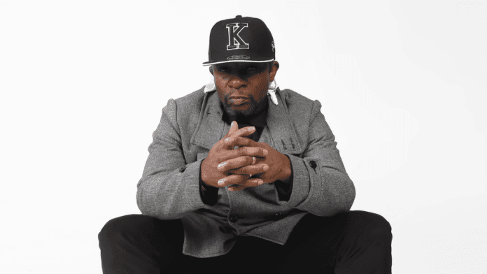 Man wearing a gray jacket and black cap sits with hands clasped, looking directly at the camera against a plain white background, channeling the vibe of Kwame Ultimate’s Global Love.