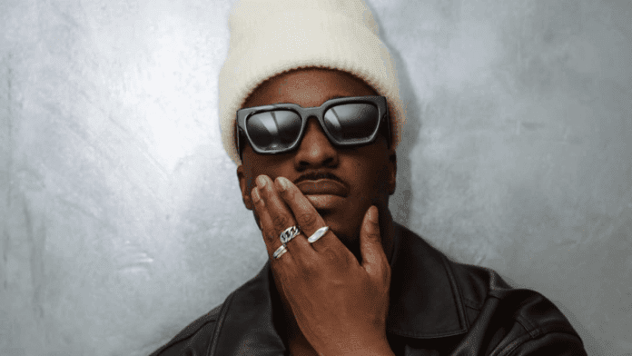 A person wearing a white beanie, black sunglasses, a black leather jacket, and multiple rings poses with one hand touching their chin against a gray background, channeling Mayn Maesby's cool style from his latest Afrobeats new single.