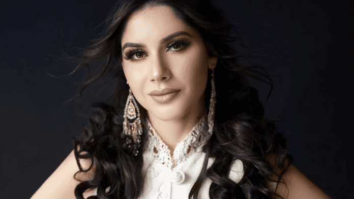 Tamara Rodríguez, in an Afro-Fusion style, poses against a dark background with long dark wavy hair, a white lace top, and ornate earrings.