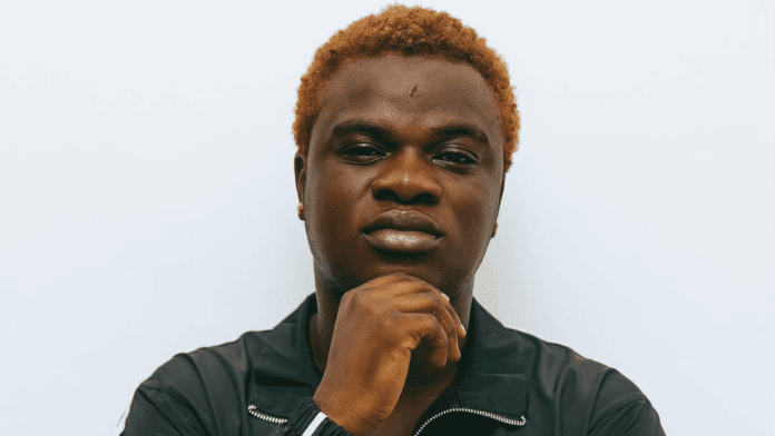 A person with short, curly, light brown hair wearing a black jacket poses against a plain white background, embodying the cool confidence of an Afro Streethop artist while resting their chin on their hand.