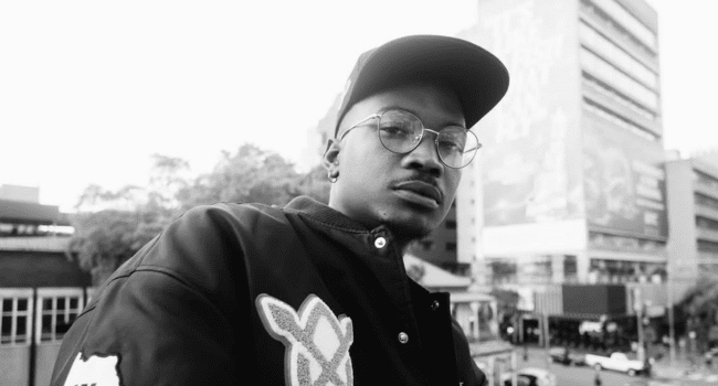 A person wearing glasses, a cap, and a jacket with patches stands outdoors in an urban area with buildings and trees in the background. The black-and-white image captures a Deluxe vibe, echoing the message: PEOPLE FORGOT TO BE PEOPLE.