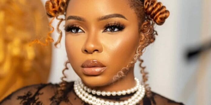 A woman with styled hair, bold eye makeup, and glossy lips wears layered pearl necklaces and a black lace outfit, channeling Yemi Alade’s signature flair—perfect for the release of her new single, "My Bebe.