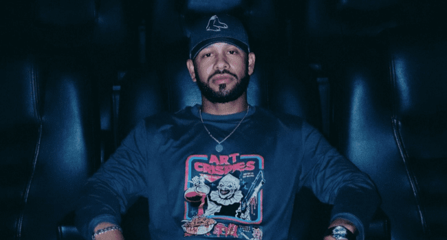 A man with a beard and mustache sits in a dark room wearing a cap and a sweatshirt that says "ART CRISES" with a graphic design on the front, channeling the style of YoungstaCPT.