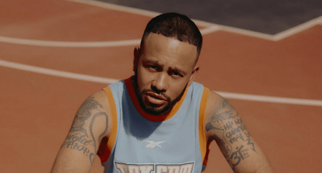 A man with tattoos, wearing a blue Reebok basketball jersey, sits on an outdoor basketball court, channeling the energy of SA Hip Hop as he looks towards the camera.