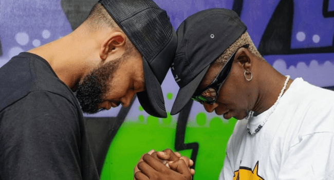 Two men stand close with their heads bowed and hands clasped together in front of a colorful graffiti wall, sharing an emotional tribute inspired by Bravo Le Roux.
