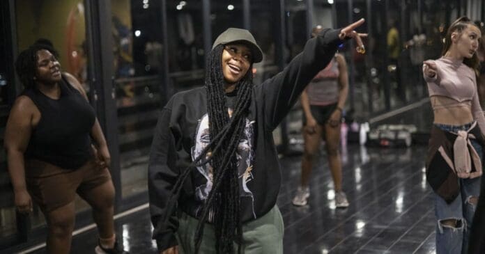 A group of people in casual workout clothes follow an amapiano dance routine in a studio with mirrored walls; one woman in front points and smiles, capturing the spirit of world dancing inspired by South Africa.