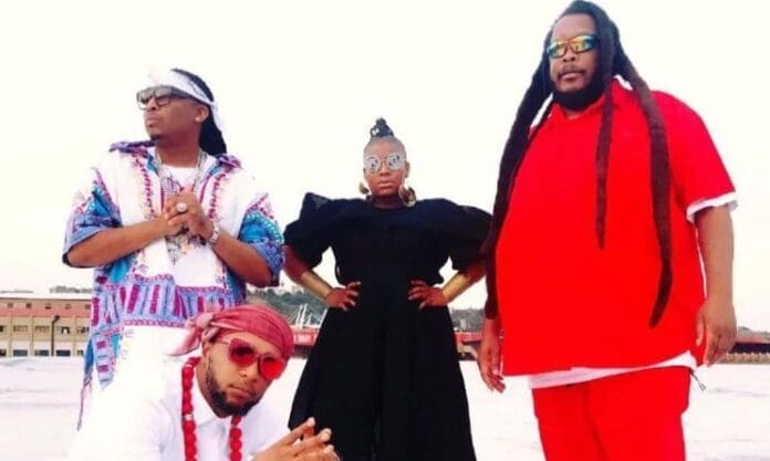 Four people pose together outdoors in colorful, distinctive outfits at the Oliver Mtukudzi International Festival, with a building and clear sky in the background.