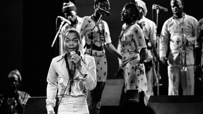 A black-and-white photo of African musician Fela Kuti singing into a microphone, with his band and backup singers performing behind him on stage—a legendary figure honored with a Grammys Lifetime Achievement Award.