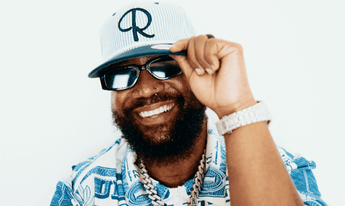 Man with a beard and sunglasses smiles, wearing a patterned blue and white shirt, a watch, and a blue-and-white baseball cap, adjusting the brim with his hand—ready for the Cassper Nyovest Fill Up concert in Bloemfontein.