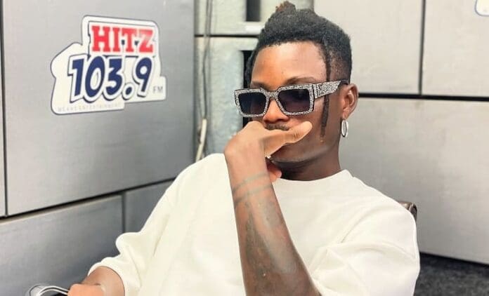 A person wearing sunglasses and a white shirt sits indoors near a wall with a "HITZ 103.9 FM" sign, resting their chin on their hand—perhaps reflecting on Chief One’s latest amapiano song making waves in Ghana.