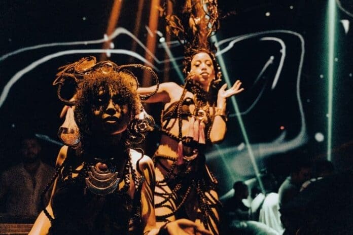 Two performers in elaborate costumes and headpieces dance under dramatic lighting with abstract patterns, evoking an Afro House vibe at a nightclub—perfect for those exploring Ibiza Spotlight or the Ibiza Virgins Guide.