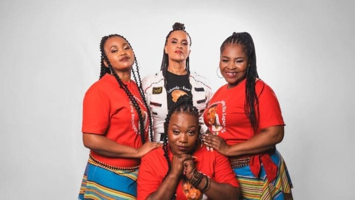 Four women pose together against a plain background; three wear matching red shirts and colorful skirts reminiscent of the Dalom Kids, icons in Music In Africa, while the fourth wears a white jacket.