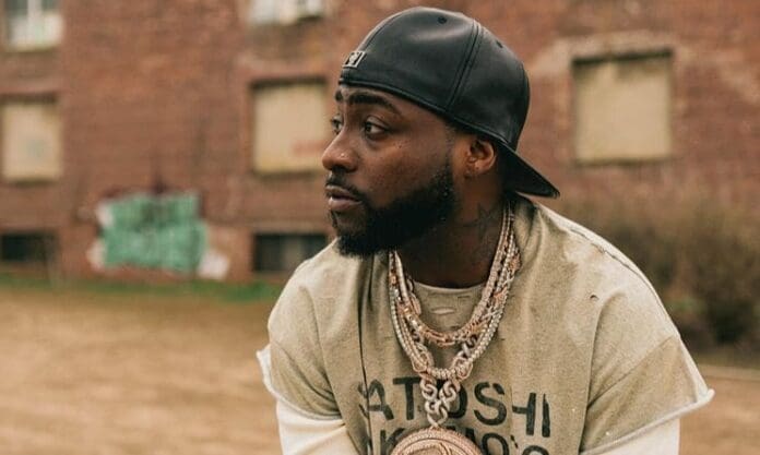 A man wearing a black cap backward and layered necklaces looks to the side outdoors at a Scorpion Kings concert in Pretoria, with a brick building in the background.