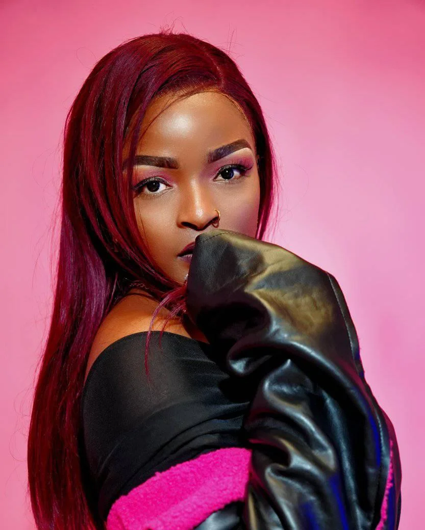 Denise, a woman with long red hair and a nose ring, poses in front of a pink background, wearing a black off-the-shoulder jacket with pink details, radiating confidence and resilience.