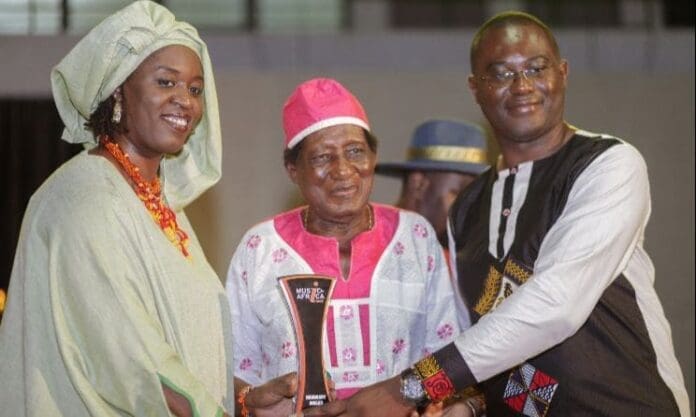 Three people pose together indoors, dressed in traditional African attire. The man in the center, known for his contributions to Ghanaian highlife, proudly holds an award trophy recognizing his impact on Music In Africa.