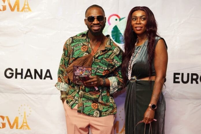 Two people pose for a photo in front of a white backdrop with "GMA Europe 2025," "Ghana," and "Europe" printed on it. One wears sunglasses, a patterned shirt, and pink pants; the other is in a green dress, capturing the spirit of music.